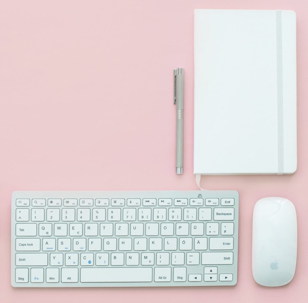 Decorative image, an Apple keyboard, mouse, silver pen and white notebook on a pink background